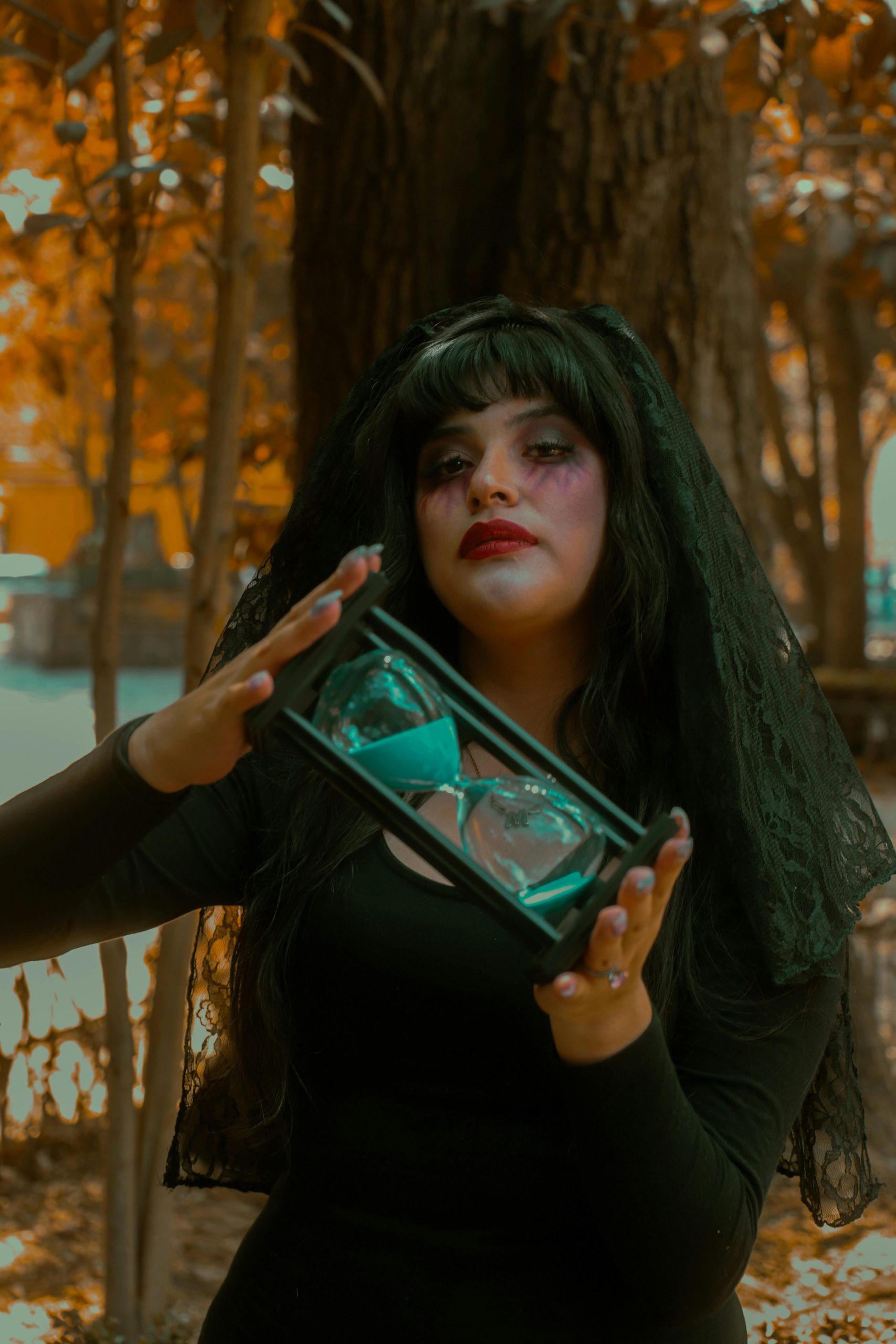 A mysterious woman in gothic attire holds an hourglass in an autumn forest setting, evoking an eerie atmosphere. Une femme mystérieuse en tenue gothique tient un sablier dans une forêt d'automne, évoquant une atmosphère étrange.
