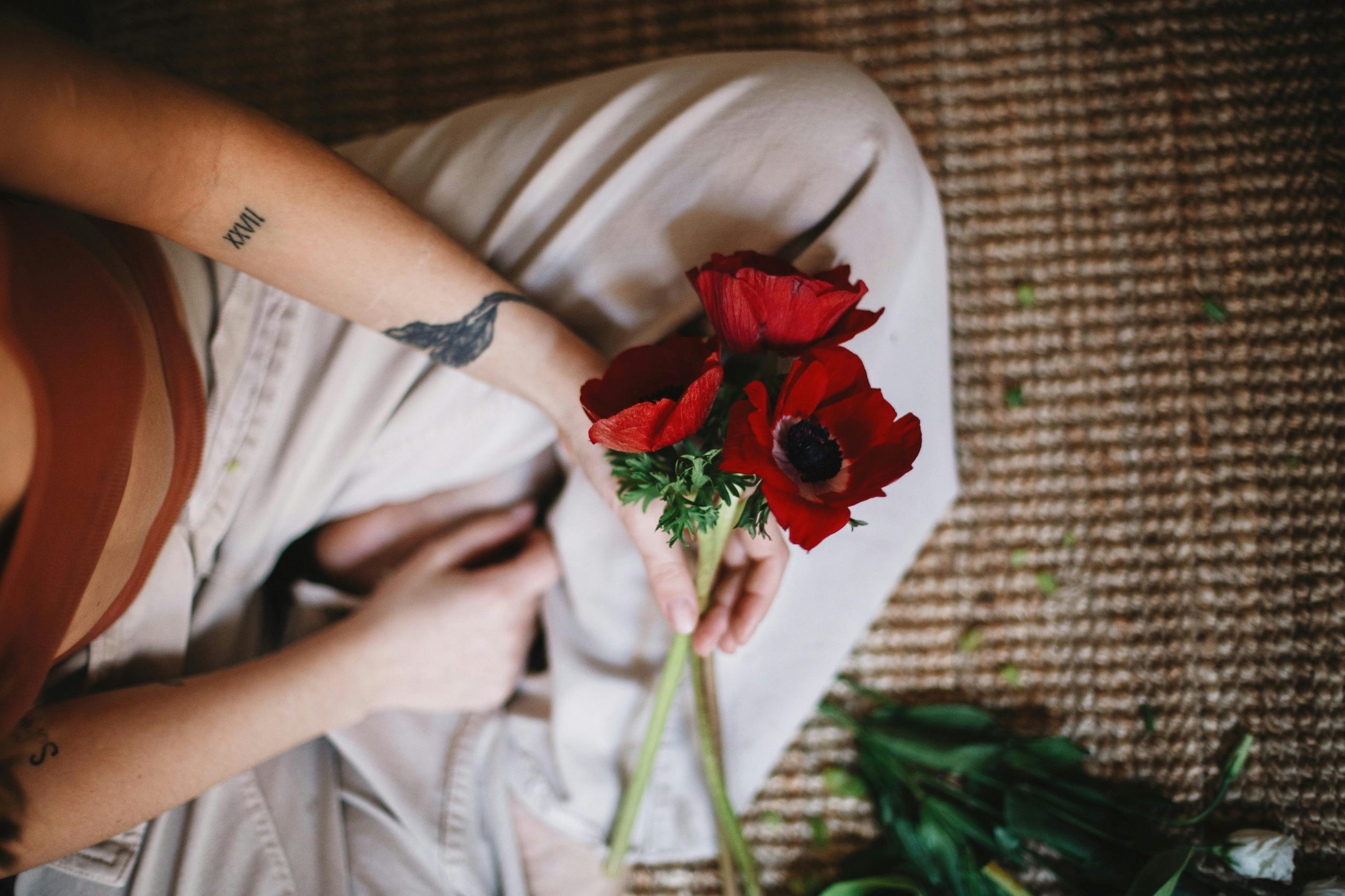 From above of crop anonymous person in casual clothes sitting with bouquet of red poppy flowers on carpet Une personne anonyme en vêtements décontractés assise avec un bouquet de coquelicots rouges sur un tapis, vue de dessus.
