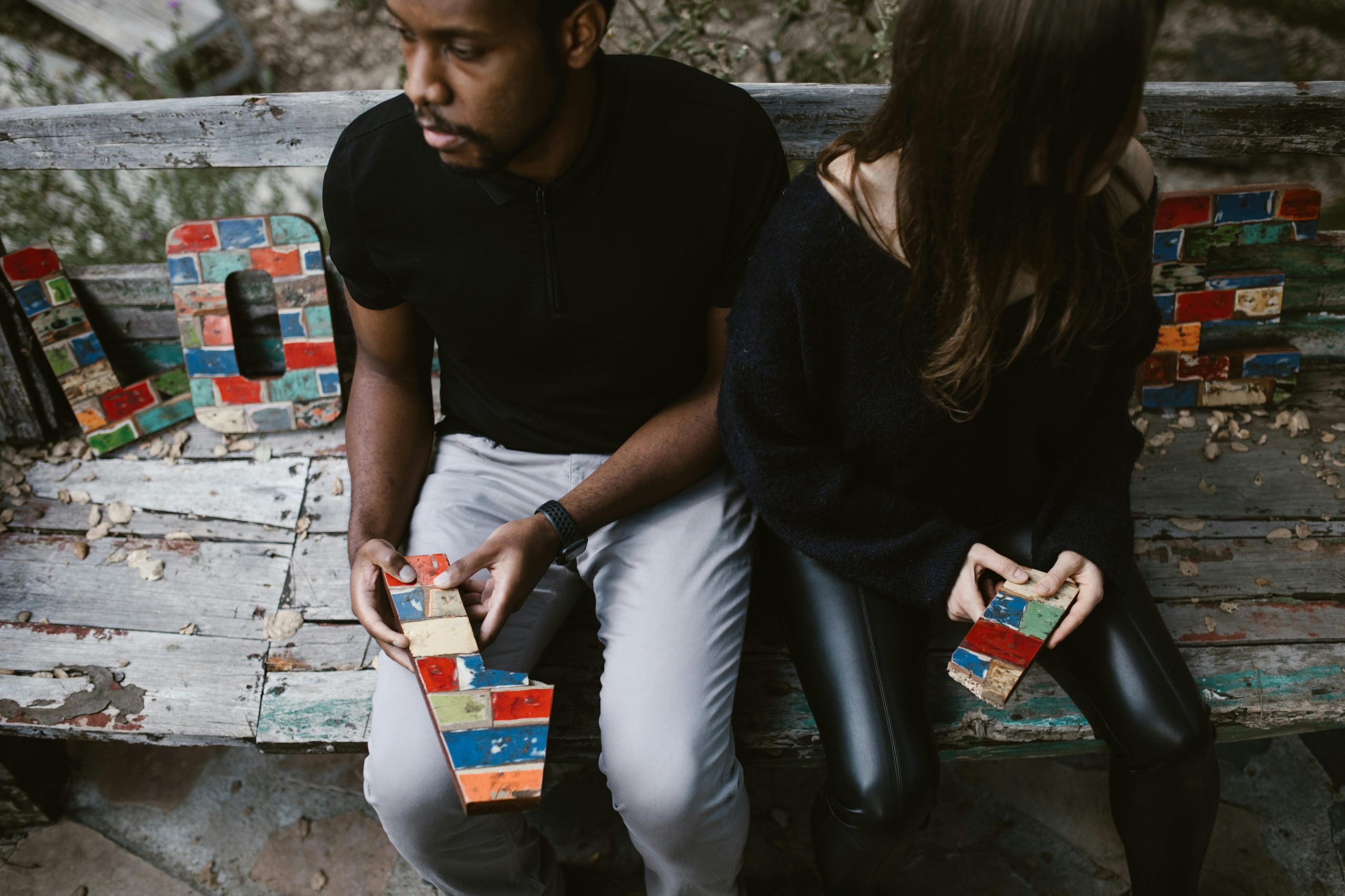 Couple assis sur un banc avec des blocs colorés formant le mot « LOVE », incarnant des émotions complexes.