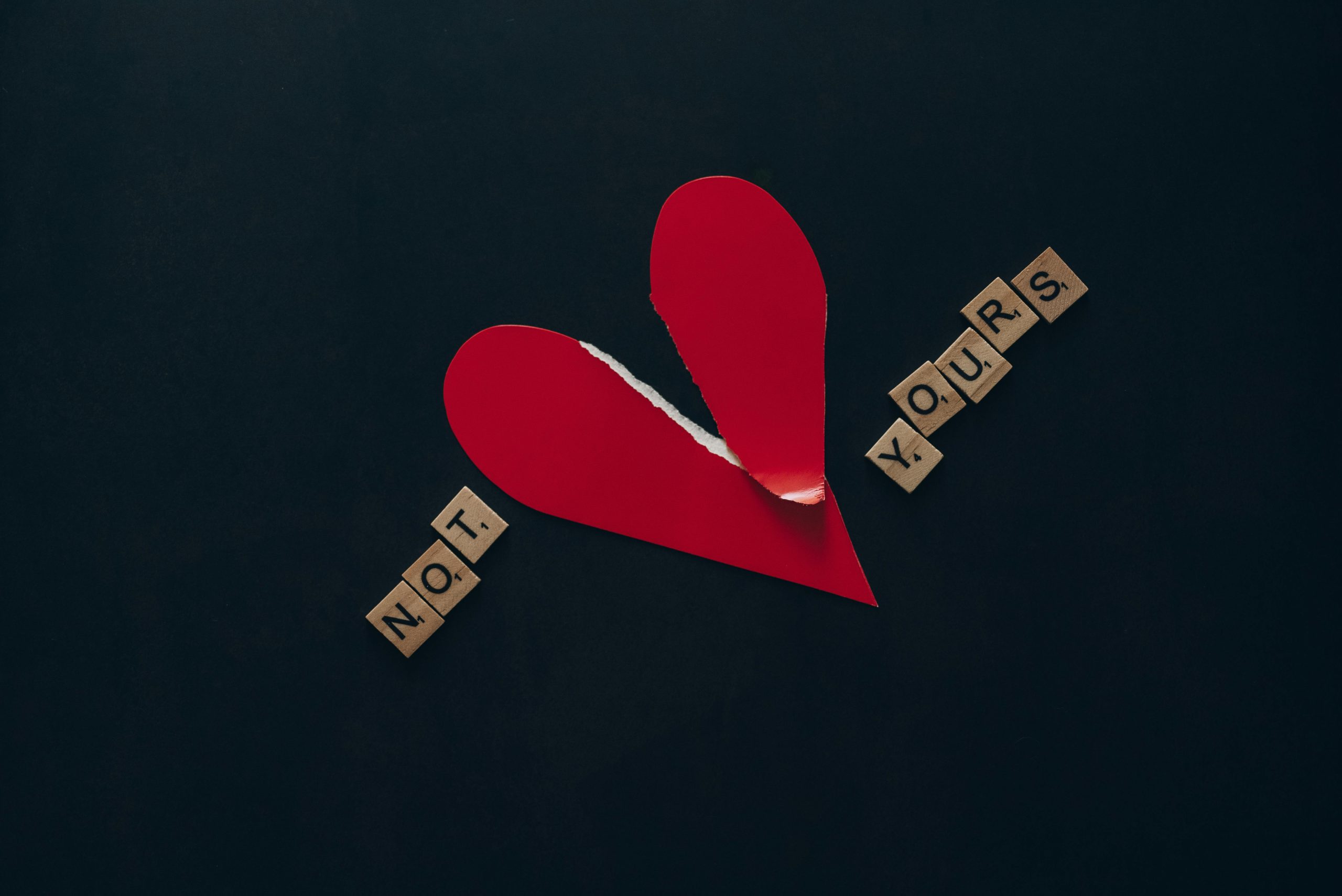Conceptual image of a torn red heart with Scrabble tiles spelling 'NOT YOURS' on a dark background. Image conceptuelle d'un cœur rouge déchiré avec des tuiles Scrabble épelant « NOT YOURS » sur un fond sombre.