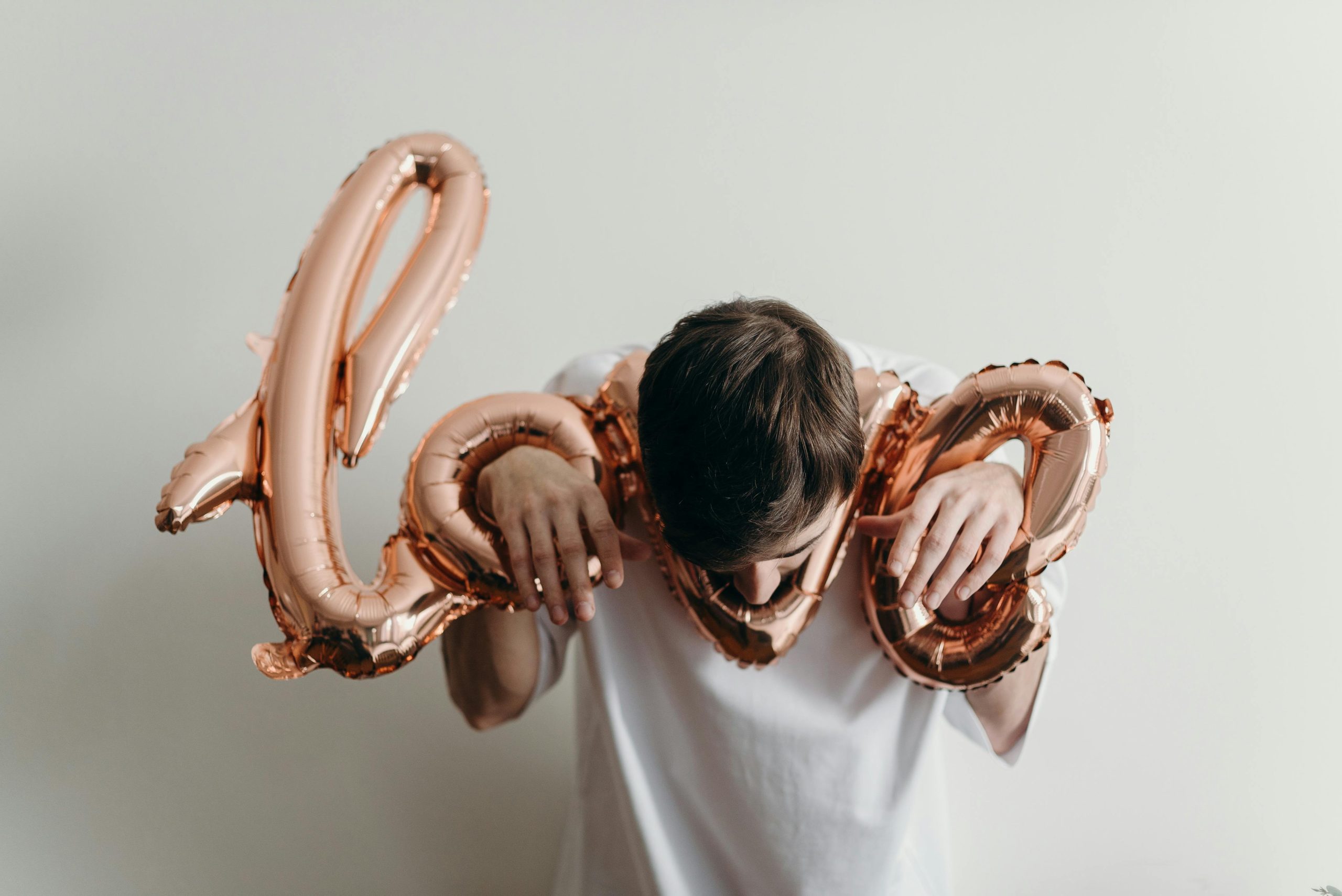 Un jeune homme en chemise blanche tenant des ballons "love" rose doré sur un fond neutre.