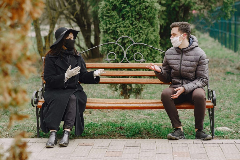 Couple sérieux non identifiable en vêtements chauds décontractés portant des gants en latex et des masques médicaux assis sur un banc dans un parc printanier en respectant la distanciation sociale