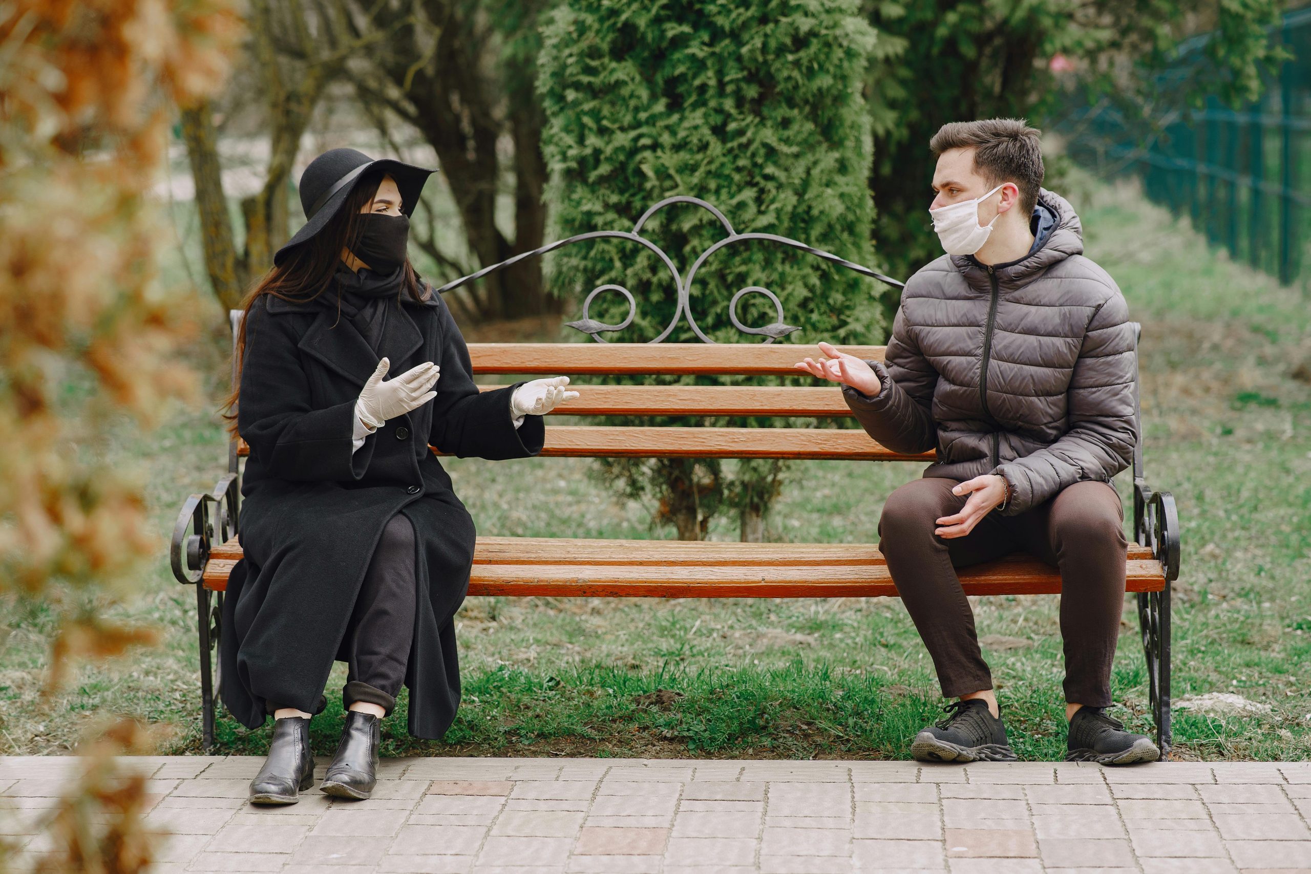 Couple sérieux non identifiable en vêtements chauds décontractés portant des gants en latex et des masques médicaux assis sur un banc dans un parc printanier en respectant la distanciation sociale