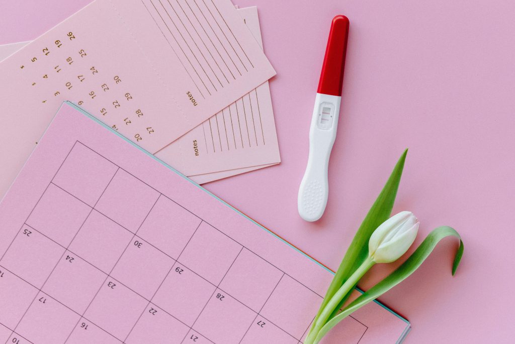 Un test de grossesse positif sur un calendrier avec une tulipe à côté sur fond rose.