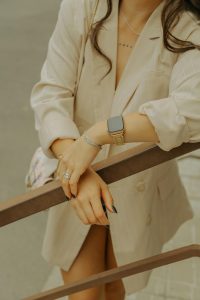 Fashionable woman in beige blazer displaying jewelry and smartwatch, leaning on a railing outdoors. Femme élégante en blazer beige affichant des bijoux et une montre connectée, s'appuyant sur une rampe à l'extérieur.
