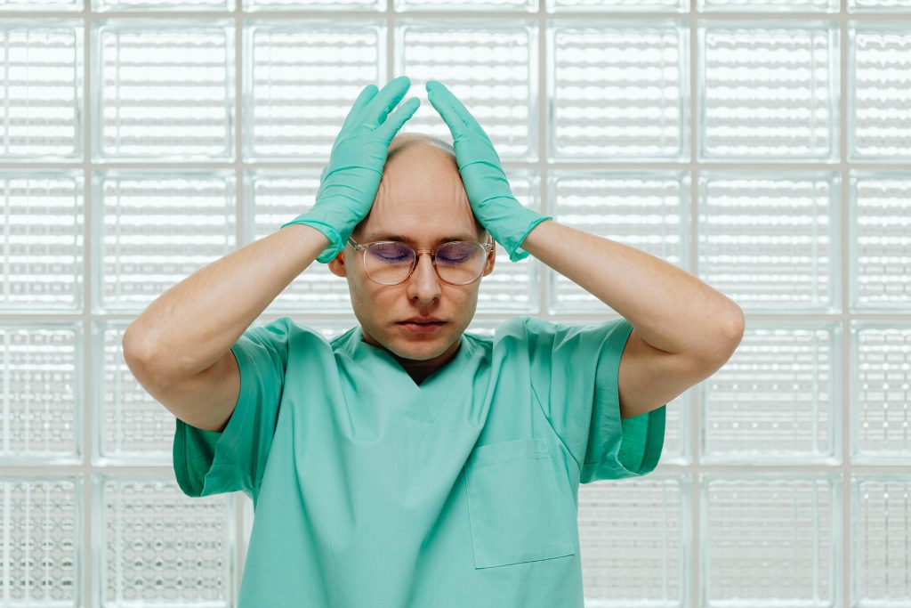 Un travailleur de la santé en blouse verte a l'air stressé, les mains sur la tête avec des lunettes.