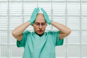 Un travailleur de la santé en blouse verte a l'air stressé, les mains sur la tête avec des lunettes.