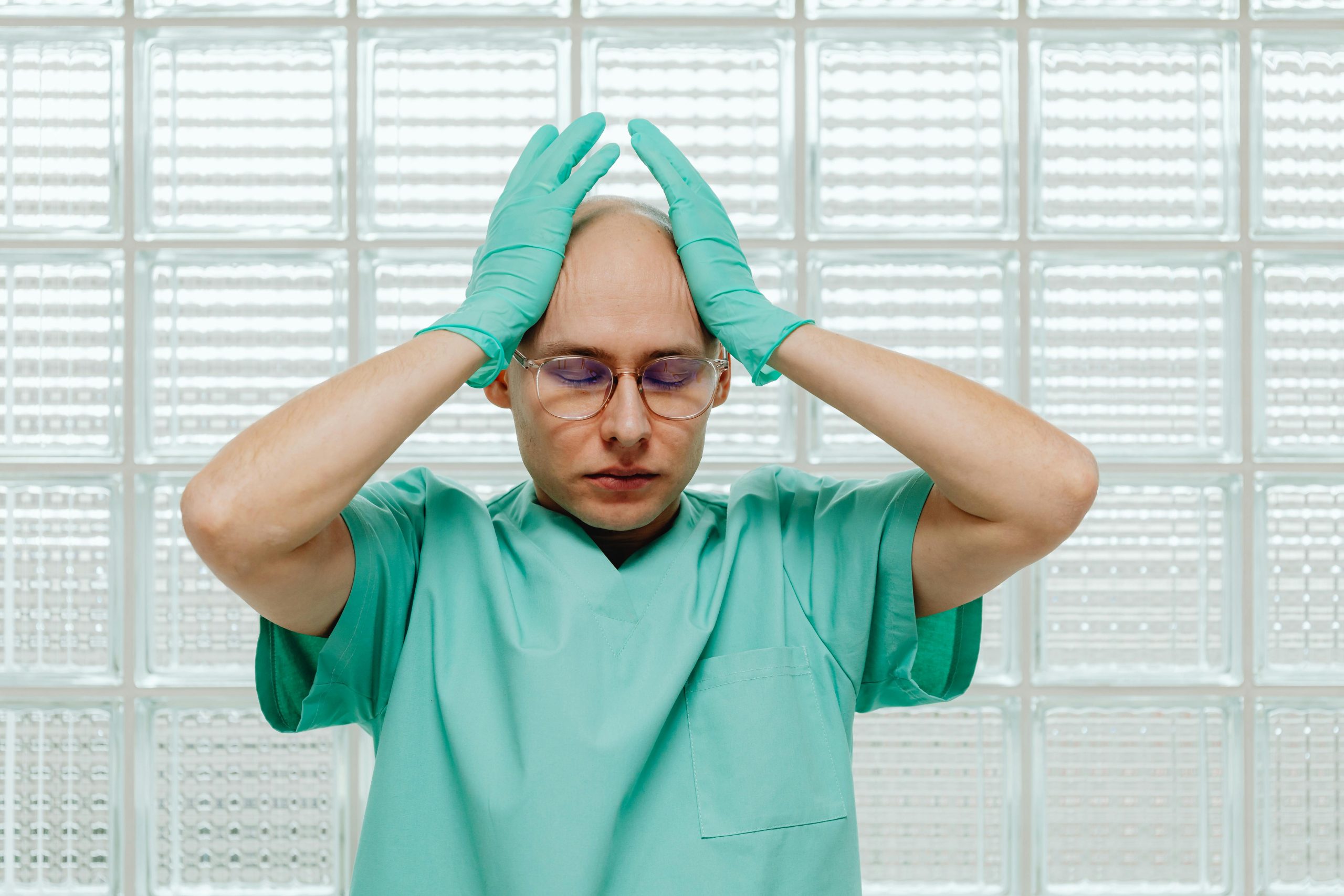 Un travailleur de la santé en blouse verte a l'air stressé, les mains sur la tête avec des lunettes.