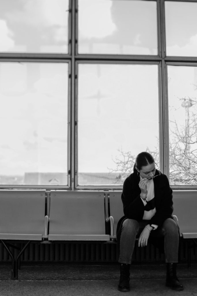 Black and white image of a woman wearing sunglasses, seated in a waiting area looking contemplative. Image en noir et blanc d'une femme portant des lunettes de soleil, assise dans une salle d'attente et regardant d'un air contemplatif.