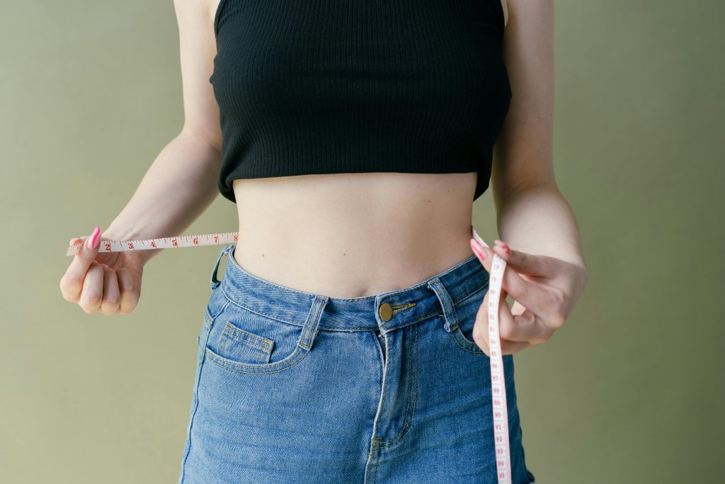 A woman in denim and crop top using measuring tape for waist check. Une femme en denim et crop top utilisant un mètre ruban pour vérifier sa taille.