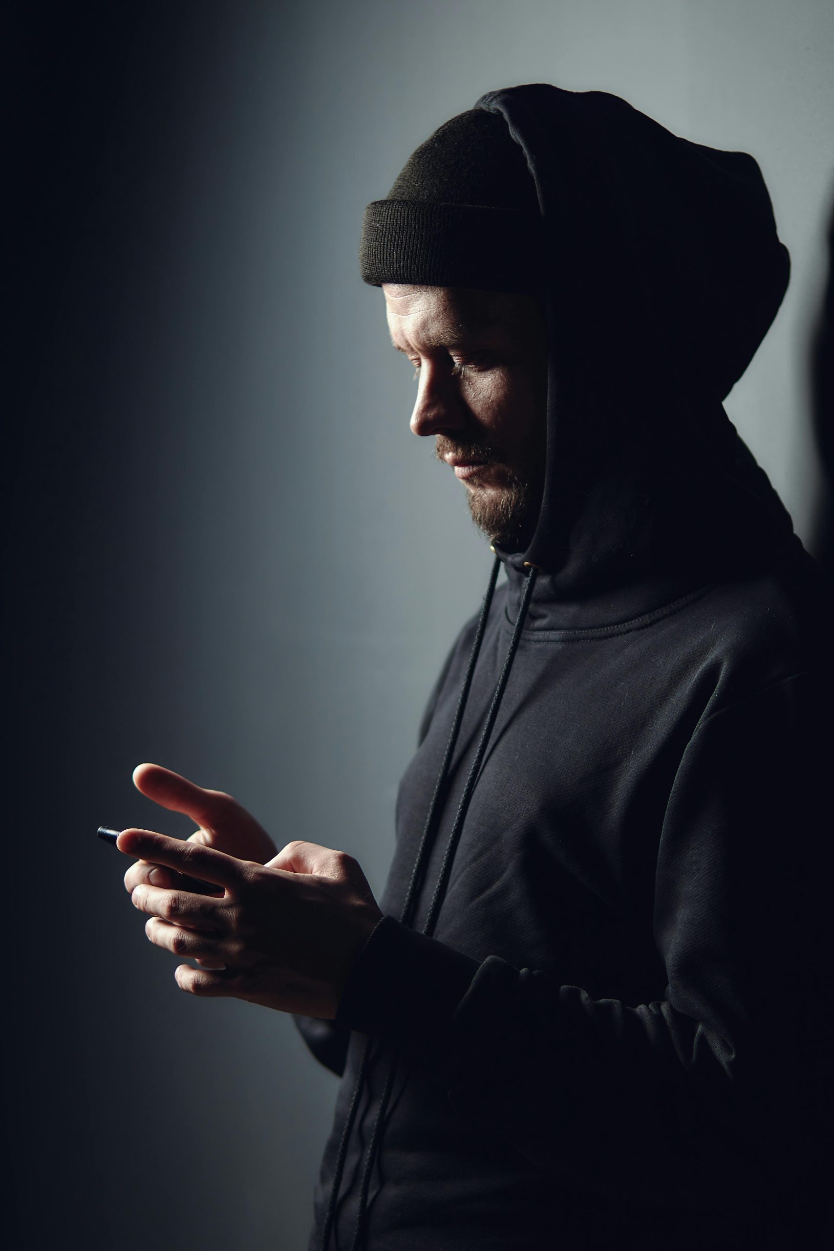 Side view portrait of a bearded man in hoodie using smartphone against dark background. Portrait en vue de profil d'un homme barbu en sweat-shirt utilisant un smartphone sur un fond sombre.