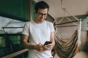 Happy man in casual t-shirt using smartphone on balcony with hammock. Homme heureux en t-shirt décontracté utilisant un smartphone sur un balcon avec un hamac.