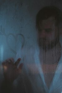A man stands behind a fogged-up shower door, drawing a heart on the glass. Un homme se tient derrière une porte de douche embuée, dessinant un cœur sur le verre.
