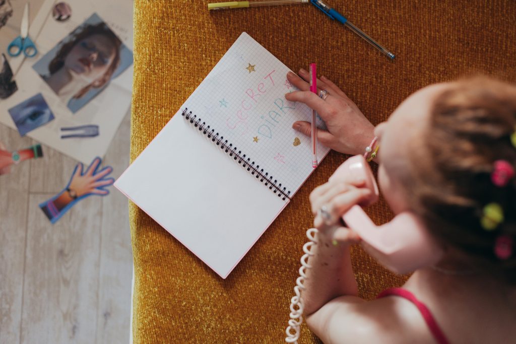 Woman writing in a diary while on a vintage pink phone, highlighting nostalgia and creativity. Une femme écrivant dans un journal intime tout en utilisant un téléphone rose vintage, mettant en évidence la nostalgie et la créativité.