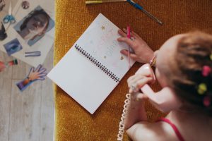 Woman writing in a diary while on a vintage pink phone, highlighting nostalgia and creativity. Une femme écrivant dans un journal intime tout en utilisant un téléphone rose vintage, mettant en évidence la nostalgie et la créativité.