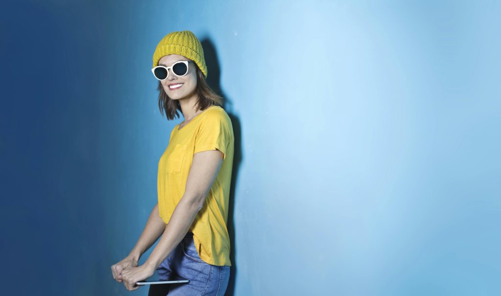 Femme souriante en tenue décontractée avec des lunettes de soleil, sur un fond bleu vibrant.