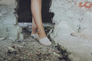 Close-up of a woman's leg wearing an ankle bracelet and espadrilles on a rocky, sandy surface. Gros plan de la jambe d'une femme portant un bracelet de cheville et des espadrilles sur une surface rocheuse et sablonneuse.