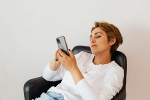Adult pensive female on casual clothes browsing social media on mobile phone while sitting on cozy leather armchair near white wall Femme adulte pensive en vêtements décontractés parcourant les réseaux sociaux sur téléphone mobile assise dans un confortable fauteuil en cuir près d'un mur blanc
