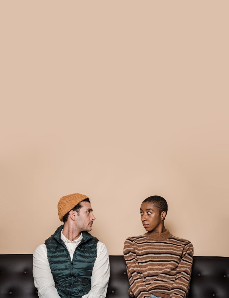 Malentendu entre un couple multiethnique ayant des problèmes relationnels, assis sur un canapé en cuir et se regardant l'un l'autre sur un fond beige.