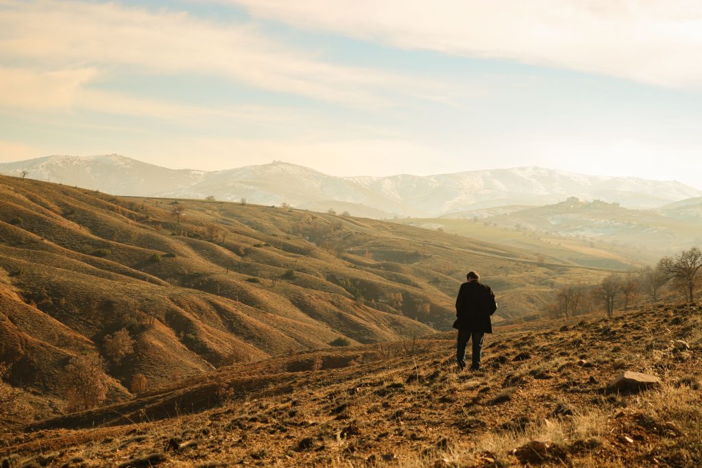 A lone person standing amidst rolling hills and distant mountains under a clear sky. Une personne solitaire debout au milieu de collines ondulantes et de montagnes lointaines sous un ciel dégagé.