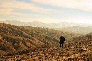 A lone person standing amidst rolling hills and distant mountains under a clear sky. Une personne solitaire debout au milieu de collines ondulantes et de montagnes lointaines sous un ciel dégagé.