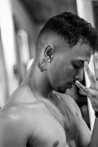 A shirtless man with a crown tattoo on his neck looks contemplative in this striking black and white portrait. Un homme torse nu avec un tatouage de couronne sur le cou a l'air contemplatif dans ce portrait en noir et blanc frappant.