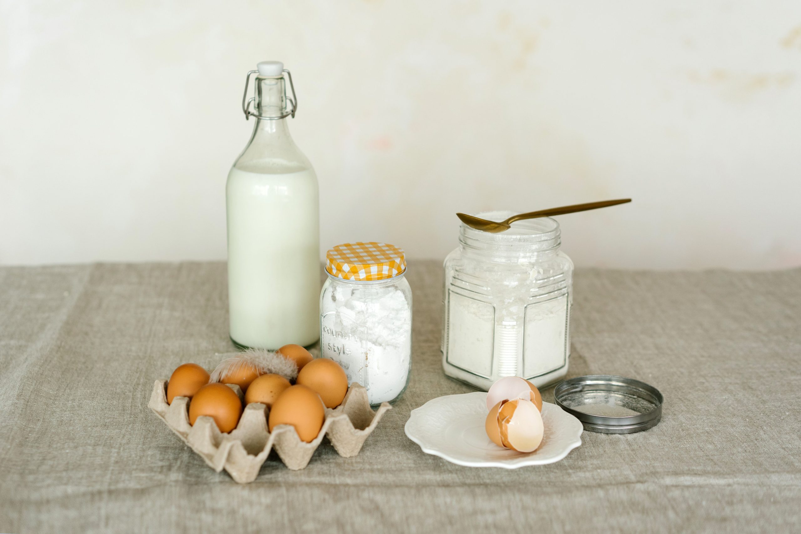 A rustic display of baking ingredients including eggs, milk bottle, and flour on a linen surface. Un affichage rustique d'ingrédients de boulangerie incluant des œufs, une bouteille de lait et de la farine sur une surface en lin.