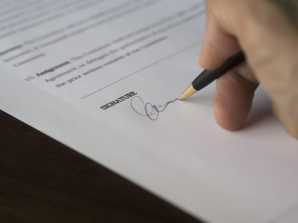 A hand signs a formal contract with a pen on a wooden desk. Une main signe un contrat officiel avec un stylo sur un bureau en bois.