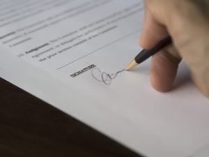 A hand signs a formal contract with a pen on a wooden desk. Une main signe un contrat officiel avec un stylo sur un bureau en bois.