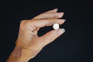 A hand wrapped in bandages holding a white pill against a dark background. Une main enveloppée de bandages tenant un comprimé blanc sur un fond sombre.