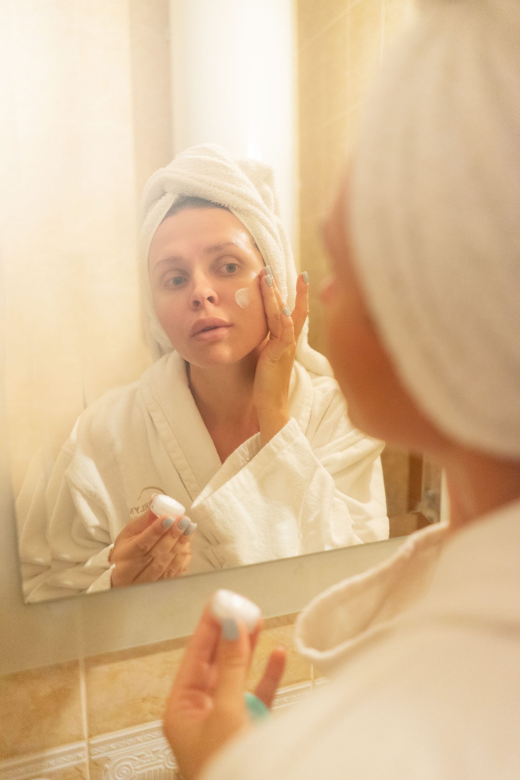 Young woman applying facial cream in front of a bathroom mirror, promoting a skincare routine. Jeune femme appliquant une crème faciale devant un miroir de salle de bain, promouvant une routine de soins de la peau.