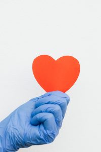 A blue gloved hand holding a red paper heart against a white background, symbolizing care and compassion. Une main gantée bleue tenant un cœur en papier rouge sur un fond blanc, symbolisant les soins et la compassion.