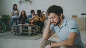 A young man sits alone looking somber while friends socialize in the background. Un jeune homme assis seul l'air morose tandis que ses amis socialisent en arrière-plan.